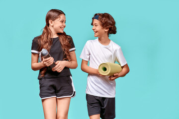 Ready for training. Two teenagers, boy and girl with yoga mat and bottle of water talking and smiling while standing isolated over blue background
