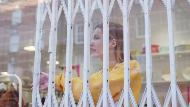 Woman running small business pushing back security grill from window and opening shop viewed from outside - shot in slow motion