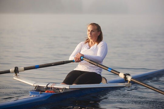 Middle aged woman rowing on lake at early morning with mist over water.