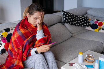 Sick young woman at home wrapped up scarf, measuring temperature with digital electronic thermometer. Health care medical concept. Disease and infection prevention. How decrease high fever