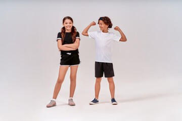 Two teenagers having workout. Happy boy showing his biceps to a cute smiling girl, standing isolated over grey background in studio