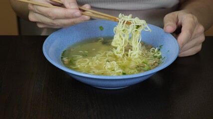 Woman sitting by the table and eating instant egg noodles with chopsticks from a ceramic dish, quick meal concept