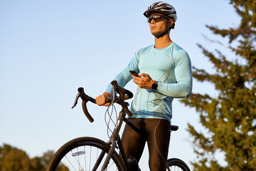 Professional male cyclist in sportswear holding smartphone and looking away, resting after ride while standing with his bike in the park on a sunny day