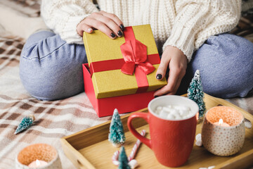 Young woman sits on bed in cozy knitted wool white sweater and holding gift-box with red gold. Hygge, New Years, Christmas. Wooden tray with a mug of cocoa with marshmallows, toy tree, candle, stars.
