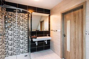Beautiful small bathroom in attic of family house. Bathroom has large glass shower,double basin with large illuminated mirror and wooden entrance door.Tiling differs a lot in terms of color/texture.