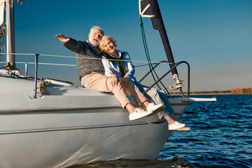 Beautiful happy elderly couple relaxing on sail boat or yacht deck floating in a calm blue sea, they looking at the horizon and enjoying amazing sunset