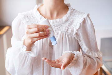 Woman in white blouse applying white antiseptic sanitizer bottle on hand against white wall. Hand hygiene, antibacterial protection. Unrecognized personality