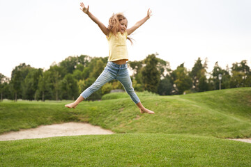 Fototapeta na wymiar Happy childhood concept. Cute little girl in casual clothes jumping and having fun while visiting park on a sunny summer day, she is smiling and feeling so excited