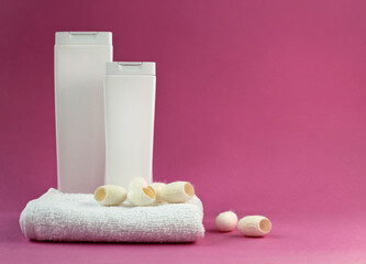 Two white bottles with shampoo, soft towel and silkworm cocoons for facial skin care on a pink...