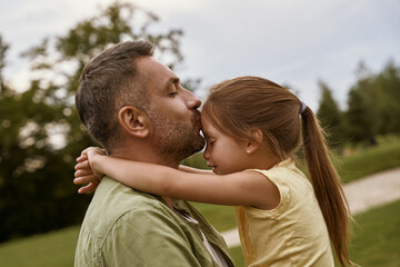 Young loving father kissing his cute little daughter in forehead while spending time together outdoors on a summer day, sitting on a green grass in park