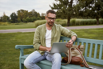 Young handsome man sitting on the wooden bench in park and using laptop, working outdoors