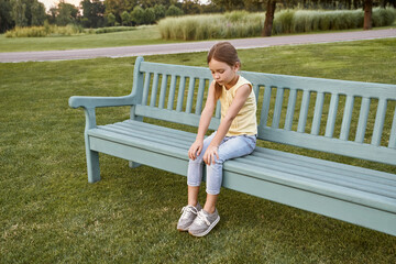 Sad little girl sitting on the bench in park on a warm day, waiting for parents outdoors