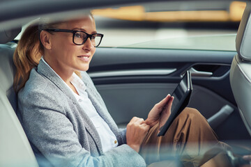 Side view of happy beautiful business woman sitting on back seat in the car and working online, she is digital tablet