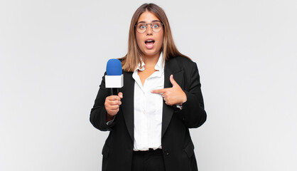 young woman reporter feeling happy, surprised and proud, pointing to self with an excited, amazed...