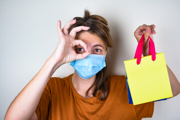 Young woman in a medical mask  holding store bags or purchases and  shows OK gesture on color...
