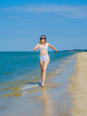 Fototapeta na wymiar A young girl runs along the sandy sea beach along the surf. Splashes of sea water. The girl is wearing a white swimsuit and sunglasses. Freedom and carelessness. Outdoor fitness. Sunny day. Copy space