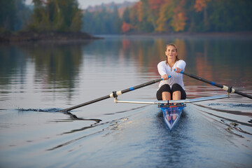 Middle aged woman rowing on a calm lake at sunrise with autumn colours in background.