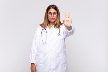 young woman physician looking serious, stern, displeased and angry showing open palm making stop...