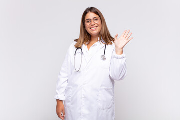 young woman physician smiling happily and cheerfully, waving hand, welcoming and greeting you, or...