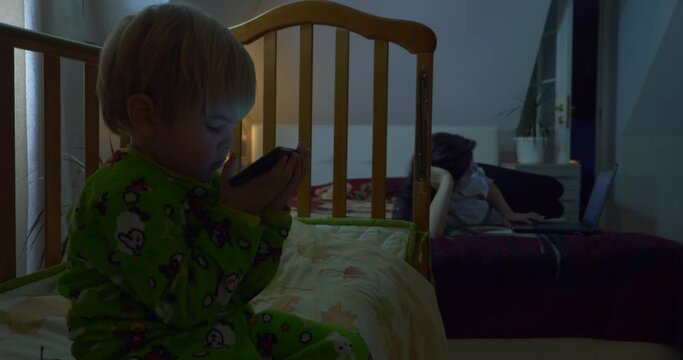 Child Watching Cartoons Videos. Mother Surfing Web Internet Browsing Social Networks on Laptop Computer. Woman Sitting on Bed in Bedroom at Home. Late Evening Night.