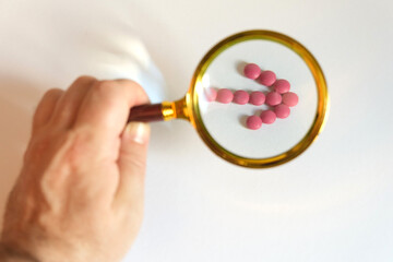 A man looks through a magnifying glass at the pink pills laid out in the form of an arrow on a white background.
