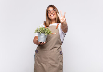 young woman gardener smiling and looking friendly, showing number three or third with hand forward,...