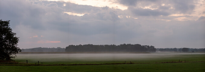 Wide panorama of a hazy dusk sunset with clouds and low hanging mist over the farmland meadow grass obscuring a tree island in the background