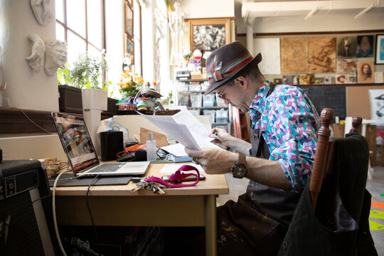 Male Artist Reviewing Paperwork At Desk In Art Studio