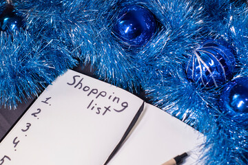 a notebook with title "Shopping list" with a pen near a blue shiny tinsel for christmas tree. new years theme.