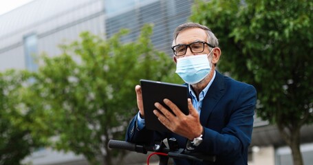 Caucasian old man wih gray hair, in glasses and medical mask sitting on bicycle and usinf tablet device. Senior male in eyeglasses tapping and scrolling on gadget computer outdoors.