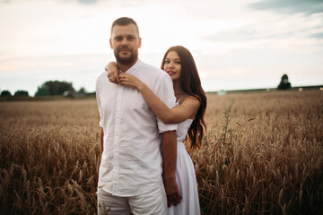 Fototapeta na wymiar Stock photo portrait of bearded boyfriend hugging his gorgeous girlfriend both in white clothes hugging in wheat field. Beautiful wheat field in the background.