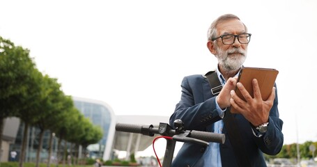 Senior gray-haired man in glasses and headphones standing at bike on street and tapping or scrolling on gadget computer. Old grandfather in eyeglasses using tablet device and browsing online outdoor.