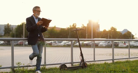 Caucasian old good-looking man in glasses standing outdoor, reading book and leaning fence. Senior grandfather read textbook or handbook. Electric scooter beside. Cars parking.