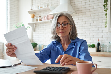 Senior mature business woman holding paper bill using calculator, old lady managing account finance, calculating money budget tax, planning banking loan debt pension payment sit at home kitchen table. - obrazy, fototapety, plakaty