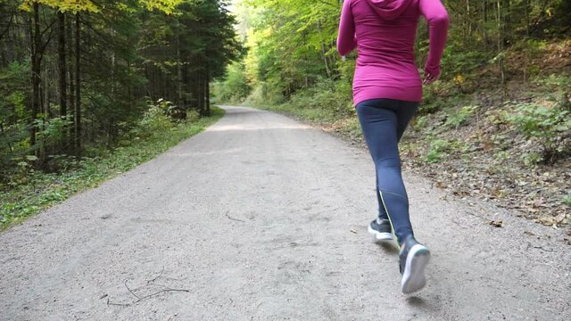 Slow motion athletic 40s woman running. Away from camera. Forest trail.