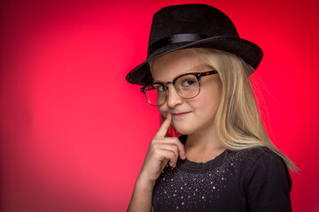 Little Girl Thinking While Wearing Glasses and Wearing a Hat Against a Red Background