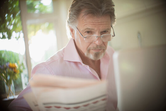 Senior man reading newspaper at laptop in sunny morning kitchen