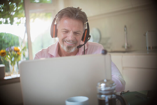 Happy senior man with headphones working at laptop in morning kitchen
