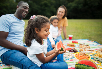Happy girl eating watermelon with family on picnic blanket in park