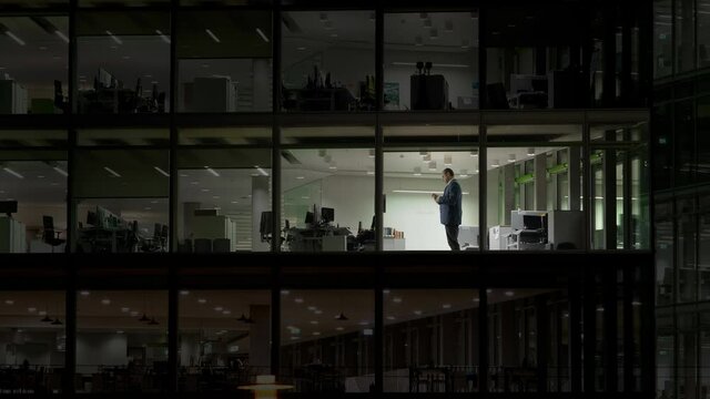 Man working late night in empty building with transparent glass window, tragic modern life without personal  happiness , work in corporate