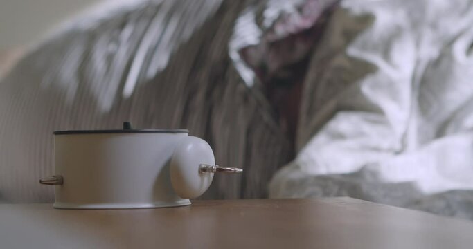 The man is woken up by an alarm clock at six in the morning. The man's hand stops the alarm clock to sleep longer. Sleeping man reluctantly wakes up to an alarm clock early in the morning.