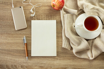 Mock up with blank notebook sheet with pen, phone, sweater and cup of hot tea on the wooden table. Cozy autumn or winter consept. 