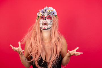 young beautiful woman wearing halloween make up over red studio background crazy and mad shouting and yelling with aggressive expression and arms raised. Frustration concept.