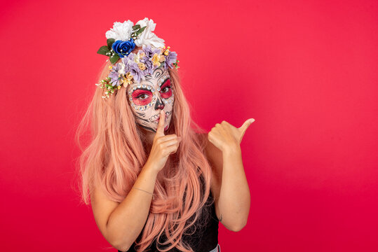 young beautiful woman wearing halloween make up over red studio background asking to be quiet with finger on lips pointing with hand to the side. Silence and secret concept.