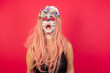 Young beautiful woman wearing halloween make up over red background angry and mad screaming frustrated and furious, shouting with anger. Rage and aggressive concept.