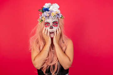 Young beautiful woman wearing halloween make up over red background, expresses excitement and thrill, keeps jaw dropped, hands on cheeks, has eyes popped out