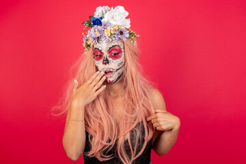 Shocked young beautiful woman wearing halloween make up over red background look surprisedly down, indicates at blank  with fore finger, . Scared male finds out price on usual shirt from luxury shop