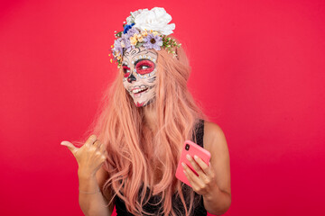 Young beautiful woman wearing halloween make up over red background using and texting with smartphone  pointing and showing with thumb up to the side with happy face smiling