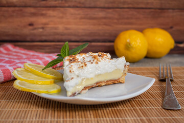 A slice of homemade lemon meringue pie on a plate, with slices of lemons, napkin completing the scene in a rustic setting and wooden planks background