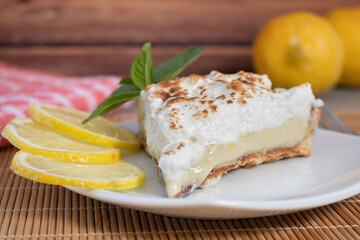 A slice of homemade lemon meringue pie on a plate, with slices of lemons, napkin completing the scene in a rustic setting and wooden planks background